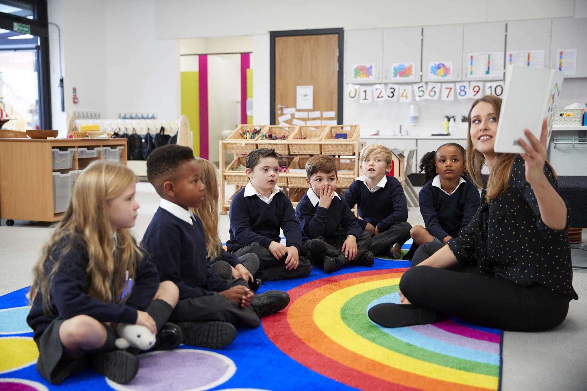 Female teacher on rainbow carpet with pupils reading a book in a classroom