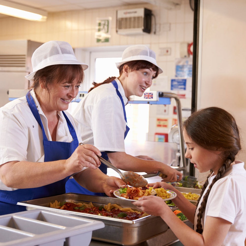 Two female school lunchtime assistants serving lunch to school children